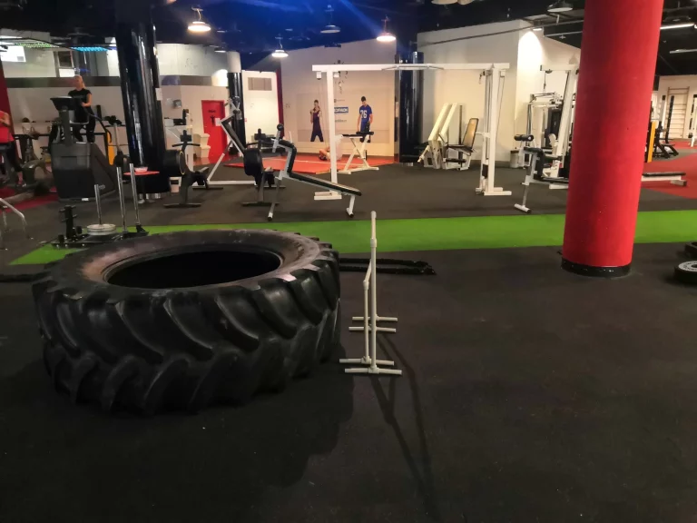 Tělocvična s různými cvičebními pomůckami, včetně velké pneumatiky na traktor, posilovacích strojů a volných závaží. Pár lidí cvičí v pozadí.